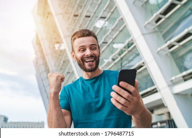 Handsome Bearded Man Showing Sincere Excitement About His Favorite Team Victory. Guy Celebrating A Bet Win In Online Sport Gambling Application On His Mobile Phone Clenching His Fist In Winner Gesture