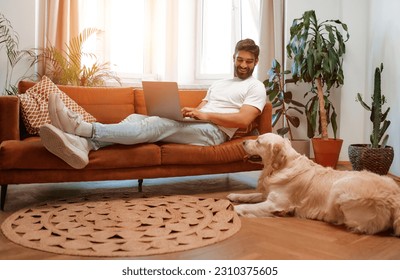 Handsome bearded man sitting on sofa by window with laptop along with labrador dog lying on floor while freelancing online, shopping or chatting in living room at home. - Powered by Shutterstock