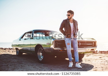 Handsome bearded man is standing near his green retro car on the beach. Vintage classic car.