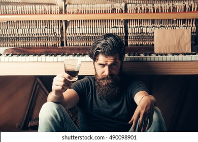 Handsome Bearded Man With Stylish Hair Mustache And Beard On Serious Face Drinking Brandy Or Whiskey From Glass Near Old Wooden Open Piano With Keyboard As Musician With Music Book, Copy Space