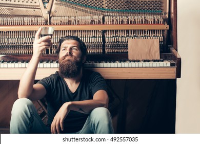 Handsome Bearded Man With Stylish Hair Mustache And Beard On Serious Face Drinking Brandy Or Whiskey From Glass Near Old Wooden Open Piano With Keyboard As Musician With Music Book, Copy Space