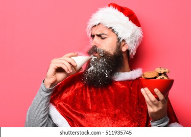 Handsome Bearded Man With Stylish Mustache And Long Snowy Beard On Serious Face Drink Milk From Glass With Chocolate Chip Cookies In Red Santa Suit On Studio Background