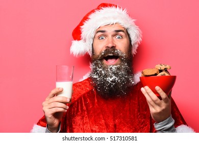 Handsome Bearded Man With Stylish Mustache And Long Snowy Beard On Smiling Face Holding Glass Of Milk With Chocolate Chip Cookies In Red Santa Suit On Studio Background