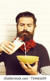 Handsome Bearded Man With Stylish Mustache And Long Beard With Red Autumn Flowers On Serious Face Pours Milk Into Yellow Bowl On White Brick Wall Background