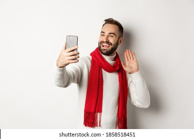 Handsome bearded man video calling friends, saying hello and waving hand at mobile phone, standing in sweater and red scarf against white background - Powered by Shutterstock