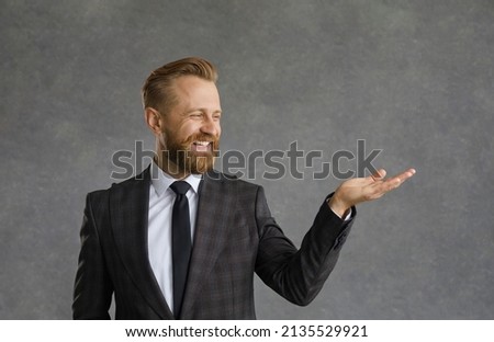 A handsome bearded man in a white shirt and black jacket with a smile demonstrates an imaginary product in the palm of his hand. The guy shows with his hand a possible product or text