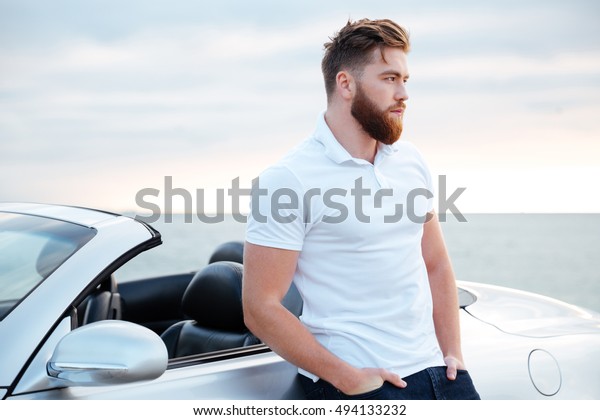 Handsome bearded man in white shirt leaning on car\
while standing on the\
beach