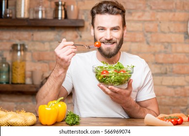 Handsome Bearded Man In White T-shirt Eating Salad With Tomatoes In The Kitchen. Healthy And Vegan Food Concept