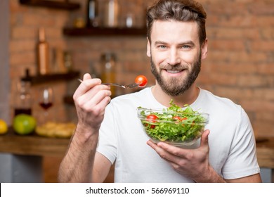 Handsome Bearded Man In White T-shirt Eating Salad With Tomatoes In The Kitchen. Healthy And Vegan Food Concept