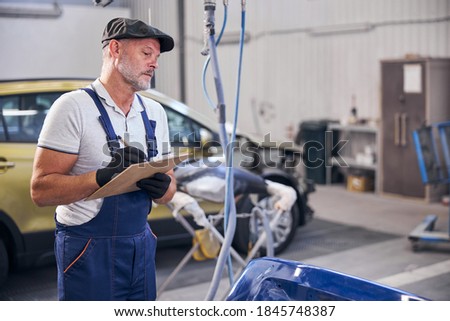 Handsome bearded man in work overalls inspecting car and taking notes while working at service station