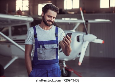 Handsome bearded mechanic in uniform is using a smart phone and smiling while standing near an aircraft in hangar