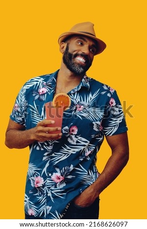 handsome bearded mid adult african american man wearing Hawaiian shirt and hat smiling with orange juice cocktail looking away at copy space on yellow background studio shot