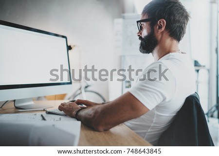 Handsome bearded professional architect wearing eye glasses working at modern loft studio-office with desktop computer.White blank display screen.Blurred background. Horizontal