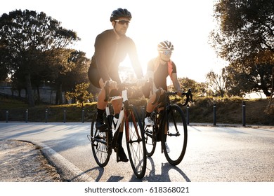 Handsome bearded professional male cyclist riding his racing bicycle in the morning together with his girlfriend, both wearing protective helmets and eyeglasses, sun shining through between them