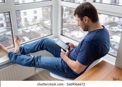 Handsome bearded relaxing man in a blue jeans and t-shirt reading an electronic book on digital device on the balcony by the window putting bare feet on a window sill in a bright modern apartment