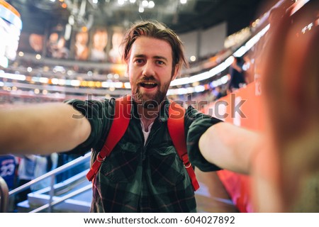 Handsome bearded supporter watching basketball game and making selfie self-portrait with smartphone at stadium