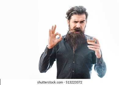 handsome bearded tough rich man with stylish hair mustache and long beard on serious face in blue fashion shirt smoking cigar holding glass of whiskey and showing ok isolated on white, copy space