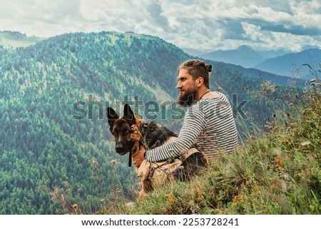 Handsome bearded Viking man with his German shepherd dog sitting on grass mountain looking at mountains in the distance on a cloudy day.