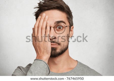 Handsome bearded young male covers eye with hands, looks through spectacles with wondering expression, afraid to see something, isolated over concrete white studio background. What is there?