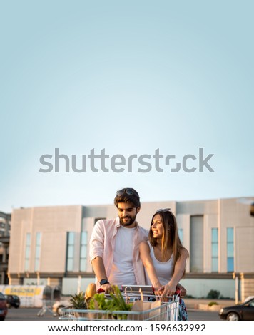 Handsome bearded young man and his wife are worried after shopping at a big market. They are in front of the shopping mall and they are pushing large carts full of groceries. Shopping. Food.