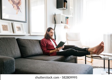 Handsome Bearded Young Man Sitting And Reading Book On Sofa At Home