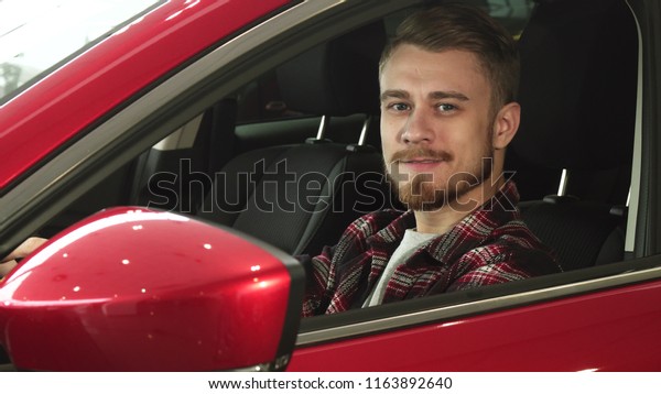 Handsome bearded young man\
sitting relaxed in his newly bought car smiling joyfully to the\
camera driving owner ownership consumerism buying shopping vehicle\
transport.