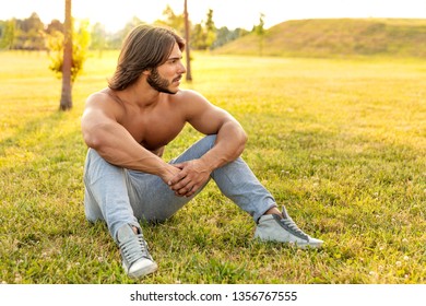 Handsome Bearded Young Man With Strong Arms And Cool Attitude Looking Away While Sitting Outdoor On The Green Grass In The Park