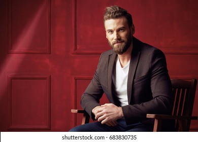 Handsome Beautiful Bearded Man, Wearing White Shirt And Brown Jacket, Sitting On Wooden Chair In Studio With Dark Red Background 