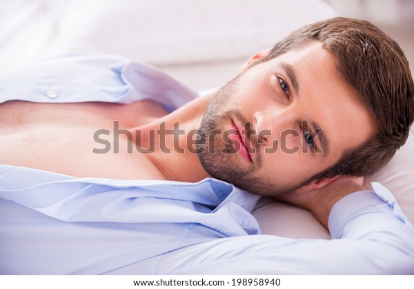Handsome in bed. Top view of handsome young man in\
unbuttoned shirt holding hand behind head and looking at camera\
while lying in bed 