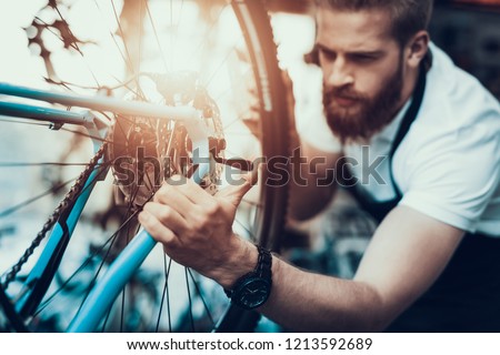 Handsome Bike Mechanic Repairs Bicycle in Workshop. Closeup Portrait of Young Bearded Man Wearing White T-Shirt Fixes Modern Cycle Transmission System. Bike Maintenance and Sport Shop Concept