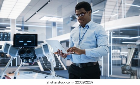 Handsome Biotechnology Engineer Using Laptop Computer, Analyzing Robotic Machine Concept in a High Tech Factory. African Scientist Manipulate and Program the Robot for Work.