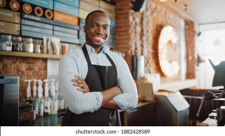 Handsome Black African American Barista with Short Hair and Beard Wearing Apron is Smiling in Coffee Shop Restaurant. Portrait of Happy Employee Behind Cozy Loft-Style Cafe Counter. - Powered by Shutterstock