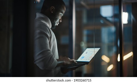 Handsome Black African American Male Is Standing In Meeting Room Behind Glass Walls With Laptop Computer In An Creative Agency. Project Manager Wearing White Jumper And Working On App User Interface.