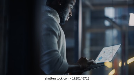 Das schöne schwarz-afrikanische Männchen steht im Konferenzraum hinter Glaswänden mit Laptop-Computer in einer Kreativagentur. Projektmanager tragen White Jumper und arbeiten an der App-Benutzeroberfläche.