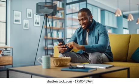 Handsome Black African American Man Using Smartphone while Sitting on a Sofa in Cozy Living Room. Freelancer Working From Home. Browsing Internet, Using Social Networks, Having Fun in Flat.