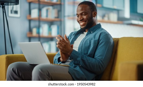 Handsome Black African American Man Working from Home and Having Video Call on Laptop Computer while Sitting on a Sofa in Living Room. He is Clapping his Hands in Celebration of the Company's Success.