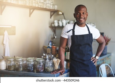 Handsome black entrepreneur stands by cafe counter lined with jars of tea while wearing dark colored apron - Powered by Shutterstock