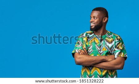 Handsome black guy in african traditional shirt looking at copy space over blue studio background, panorama. Cheerful african american young man staring at empty space for text or advertisement