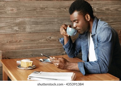 Handsome Black Male In Casual Wear Checking Email Or Reading World News On Digital Tablet Computer Drinking Morning Cappuccino, Sitting At The Wooden Table At A Cozy Cafe. Connection And Technology