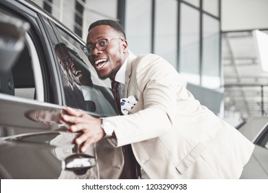 Handsome Black Man In Dealership Is Hugging His New Car And Smiling.