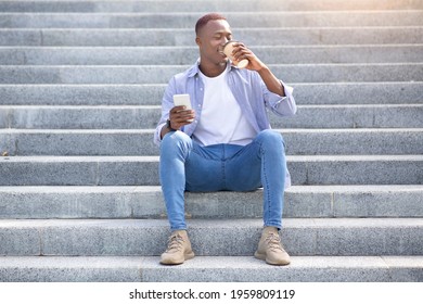 Handsome Black Man Drinking Takeaway Coffee And Using Cellphone While Sitting On Stairs Outside. Young African American Guy With Hot Drink Making Online Search On Mobile Device