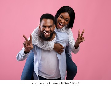 Handsome black man giving piggyback ride to his beloved woman, gesturing peace or victory on pink studio background. Young African American couple having fun together on Valentine's Day