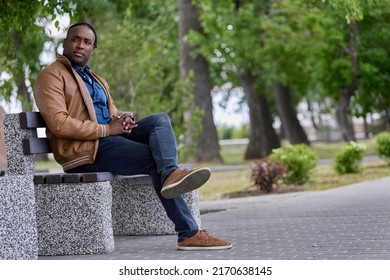 Handsome Black Man Sits On A Bench In The Park Waiting For Another Person. An African-American Man Sat Down On A Bench In An Alley In A Park, Resting From A Long Road.