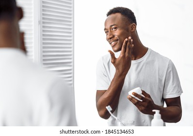 Handsome Black Man Using Facial Cream Moisturizing And Caring For Face Skin Standing Near Mirror In Bathroom Indoors In The Morning. Male Hygiene, Seflcare And Skincare Concept. Selective Focus