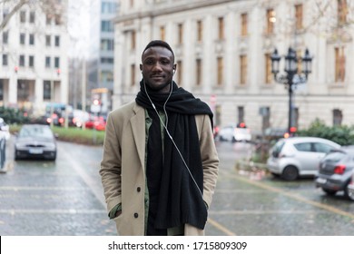 Handsome Black Real Life Model Wearing Beige Coat With Hands In Pockets In City. Looking At Camera. 