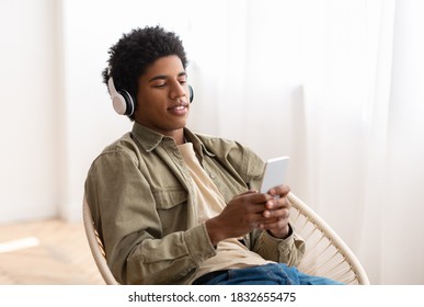 Handsome Black Teen In Headphones Choosing Musical Playlist On Smartphone At Home. Attractive African American Guy Enjoying Great Song, Listening To Audiobook Or Radio, Indoors