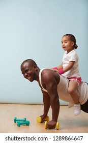 Handsome Black Young Father Is Doing Push Ups On Dumbbells With His Cute Little Daughter Riding Him. They Both Enjoy It Big Time. Indoors.