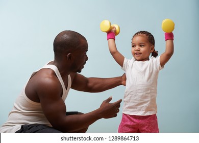 Handsome black young father instructing his cute little daughter on how to exercise with dumbbells against pale blue background. Lifting dumbbells overhead. She smiles and looks at him for approval. - Powered by Shutterstock