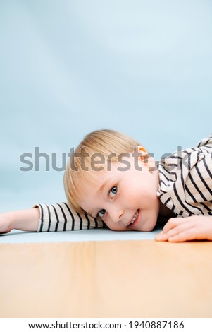 Handsome blond boy lying on the floor, playing cute. He's wearing striped long-sleeve shirt. Studio shot. Over blue background. Looking at the camera.