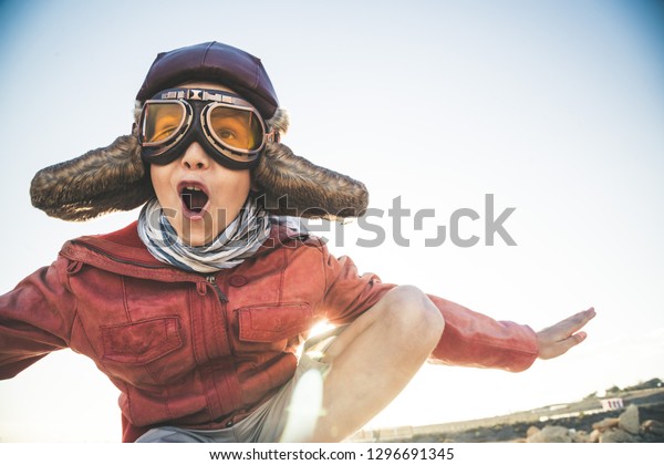 Handsome blond boy plays happy and joyful pretending\
to take off his flight disguised as a vintage aviation pilot with\
hat, leather jacket yellow eyewear mask and fluttering foulard\
Screaming with joy